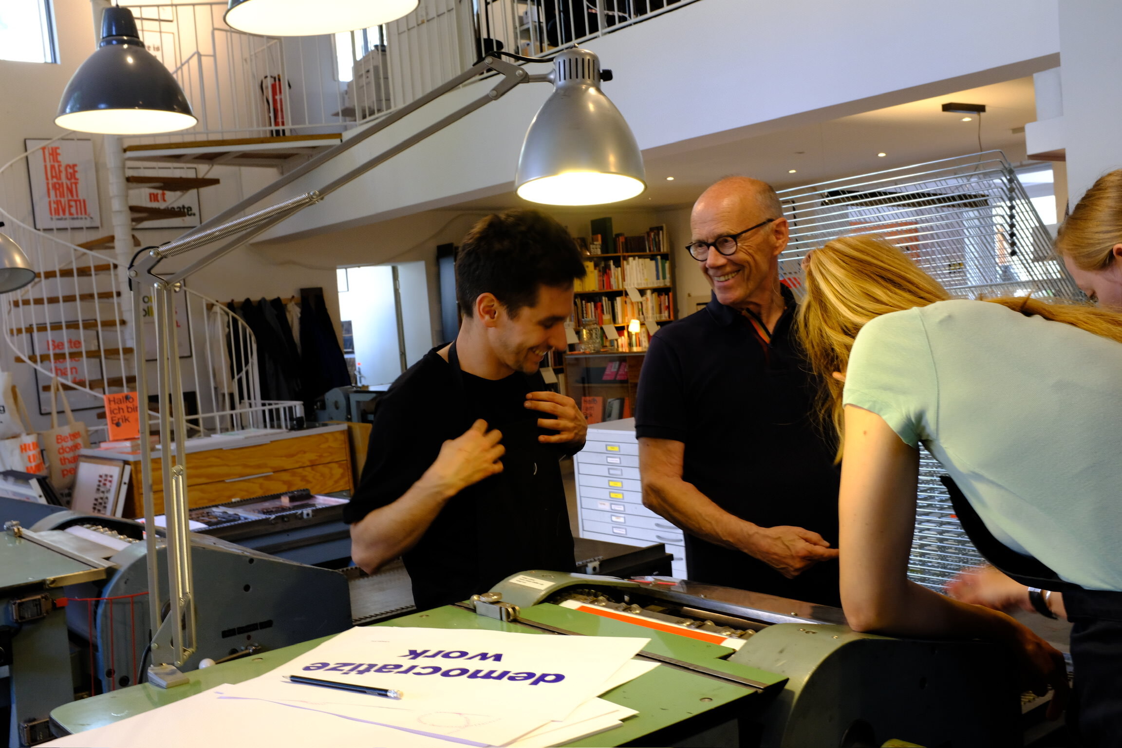 Photo of four people standing around a letterpress machine, laughing