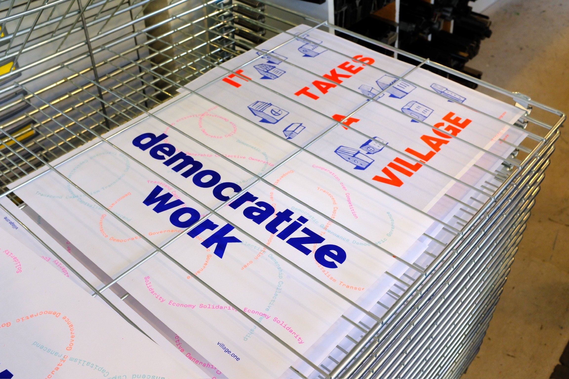 Photo of two printed posters in a drying rack, saying “democratize work” and “it takes a village”