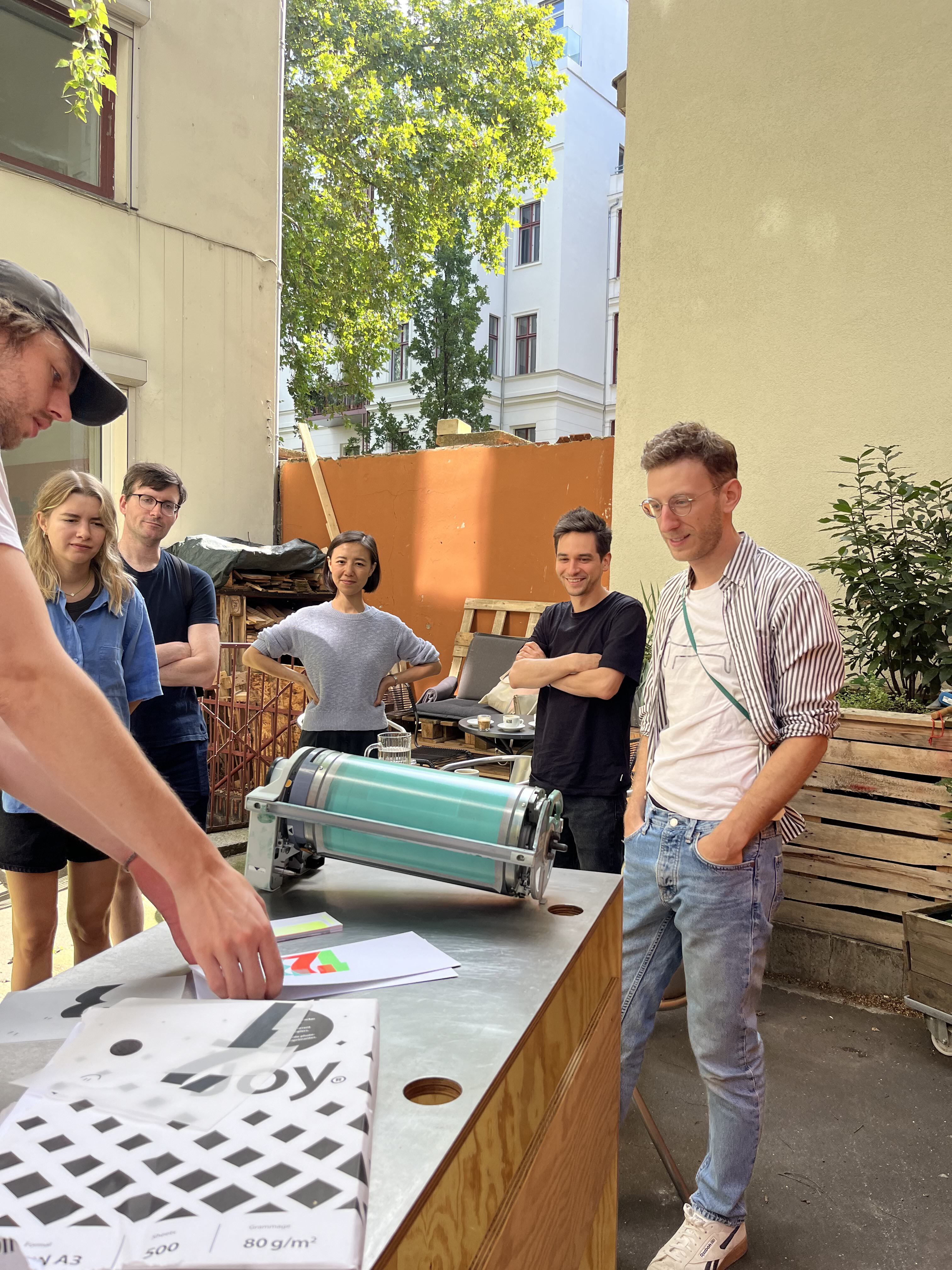 Photo of a group of people standing outside around a table, looking at some printed material and a paint roll for a riso printer