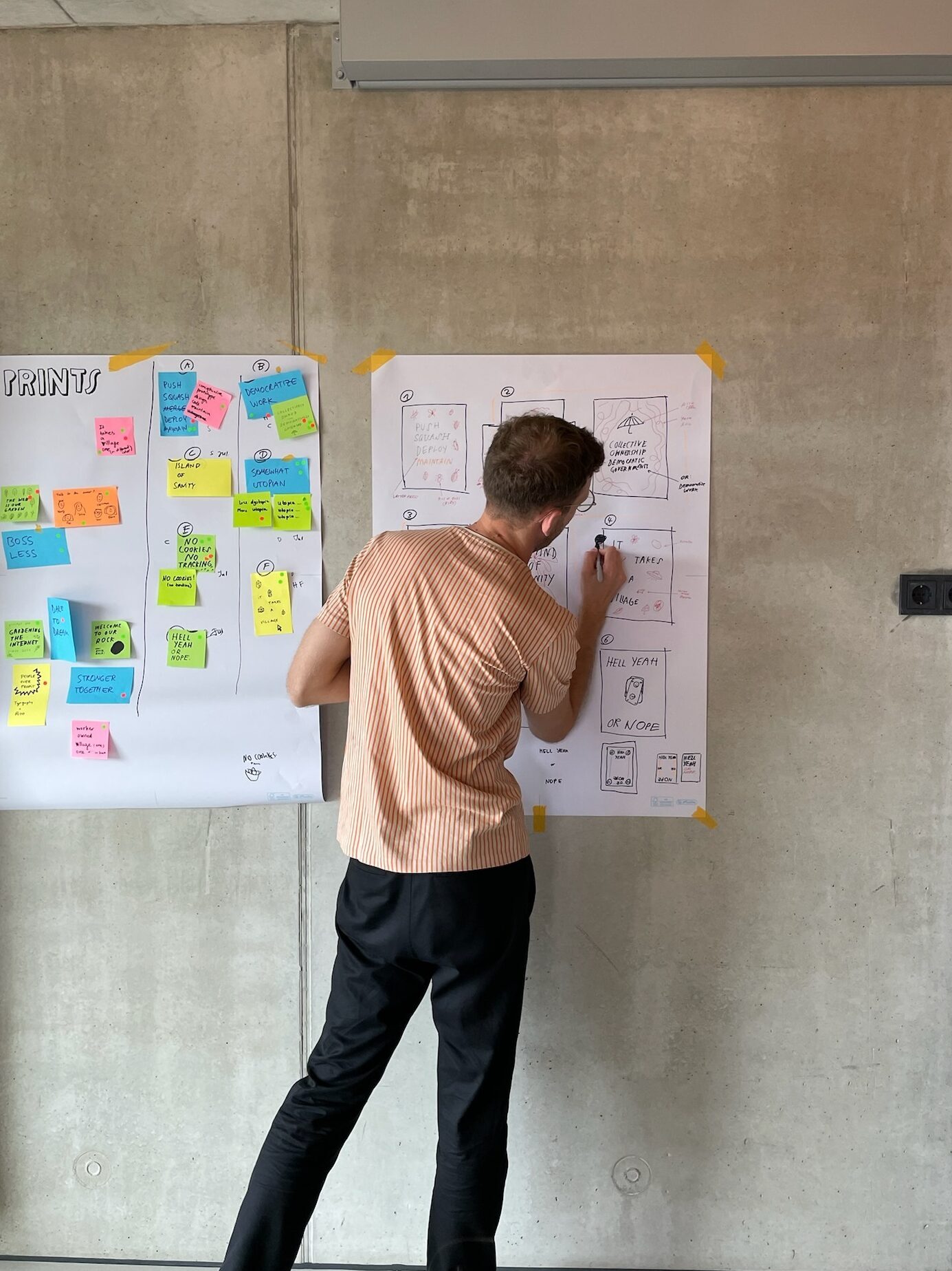 Photo of a dude standing in front of a concrete wall, with a poster in front of him, pen in his hand, scribbling ideas onto the paper