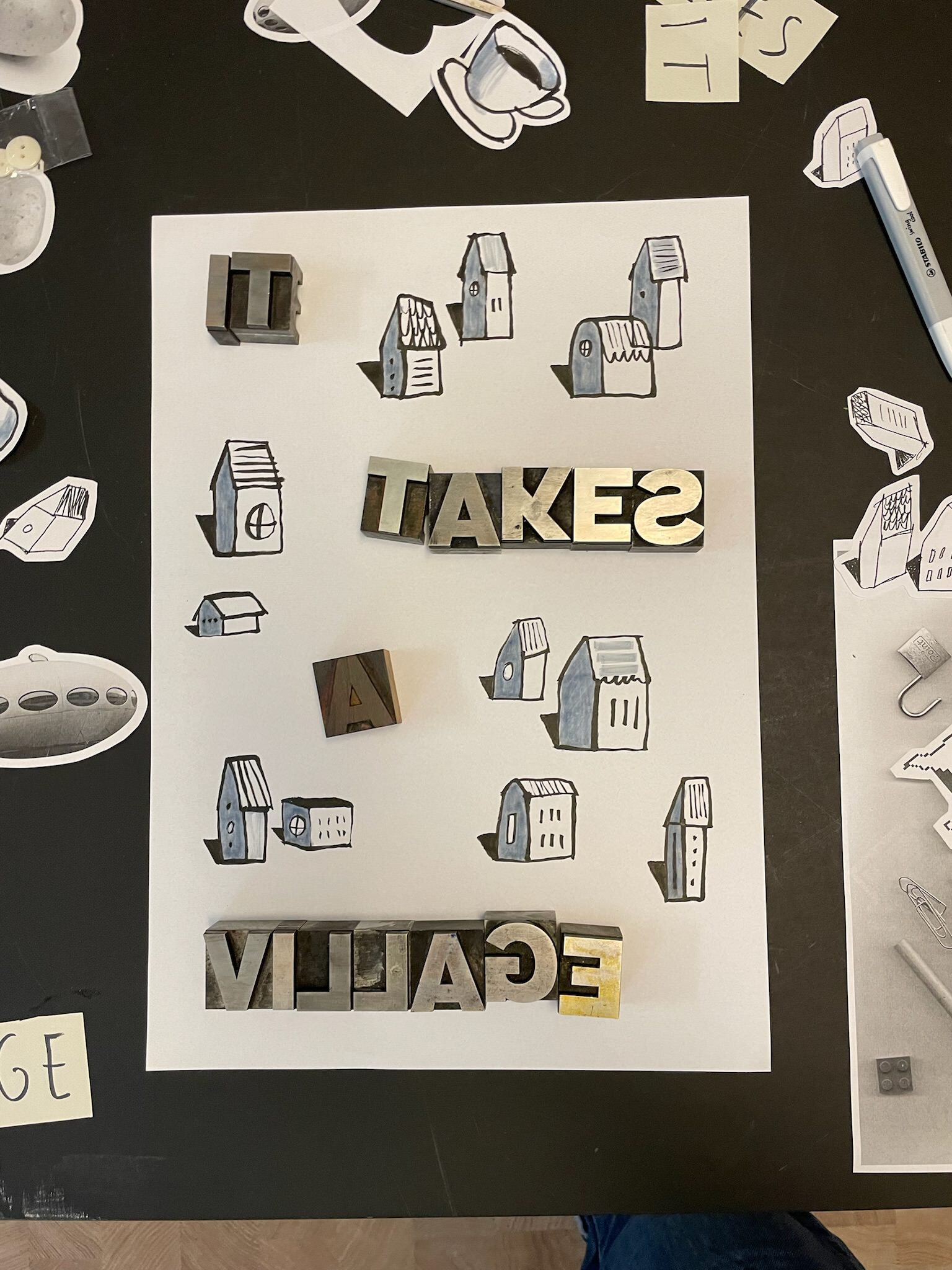 Photo of a poster with lots of little drawn houses and metal type arranged on top, spelling out the words “It takes a village”