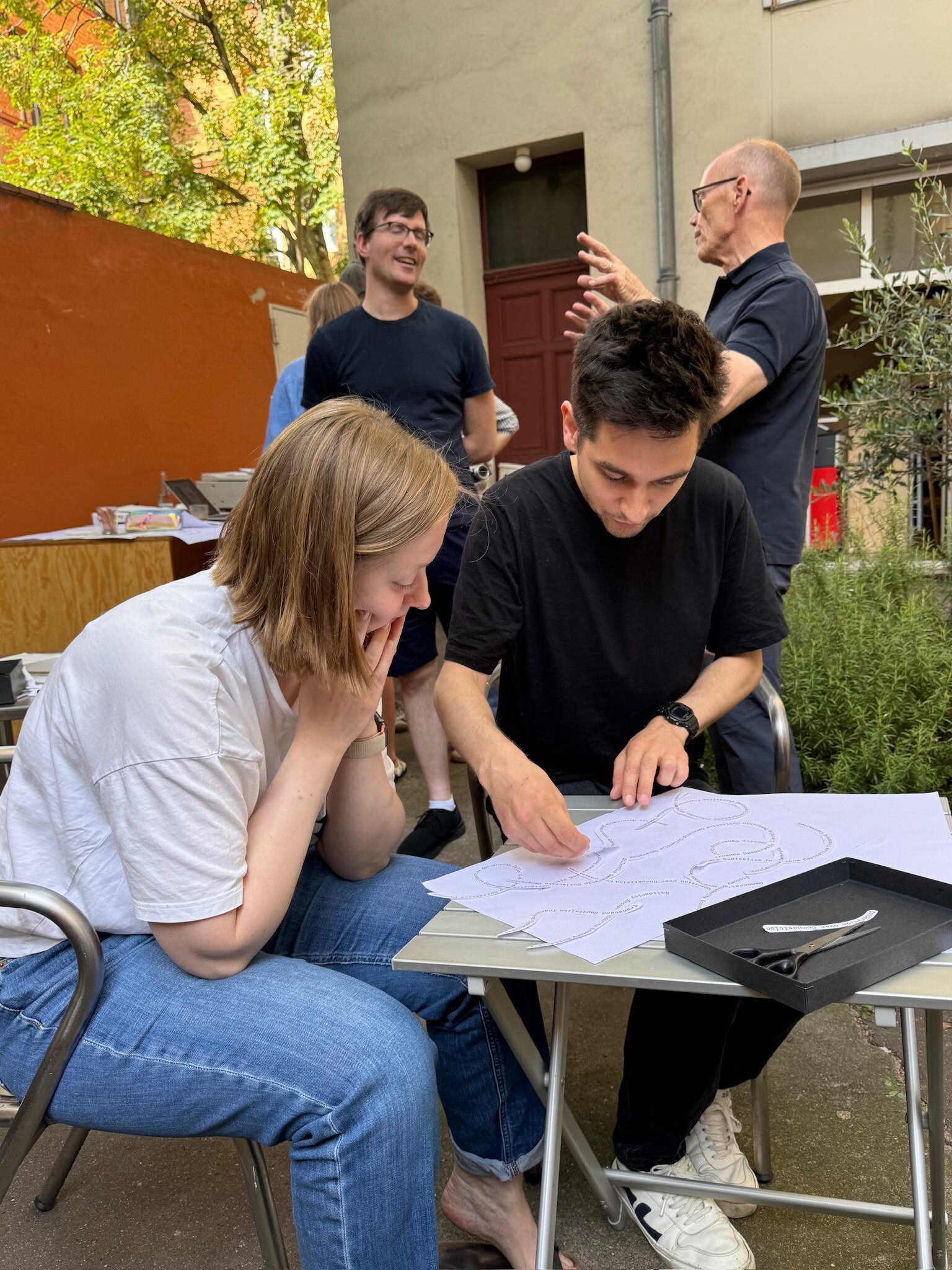 Photo of two people sitting on a table arranging pieces of paper, with more people in the background discussing