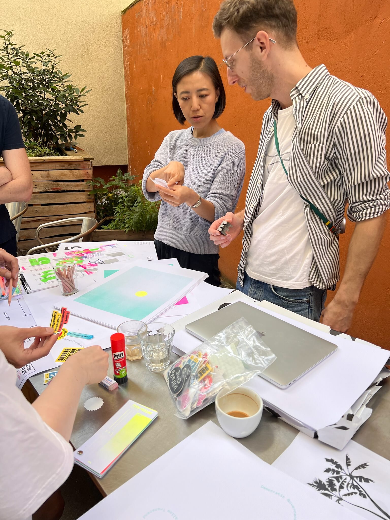 Photo of two people standing behind a table with lots of crafting material on top, discussing a piece of paper one person holds in their hand