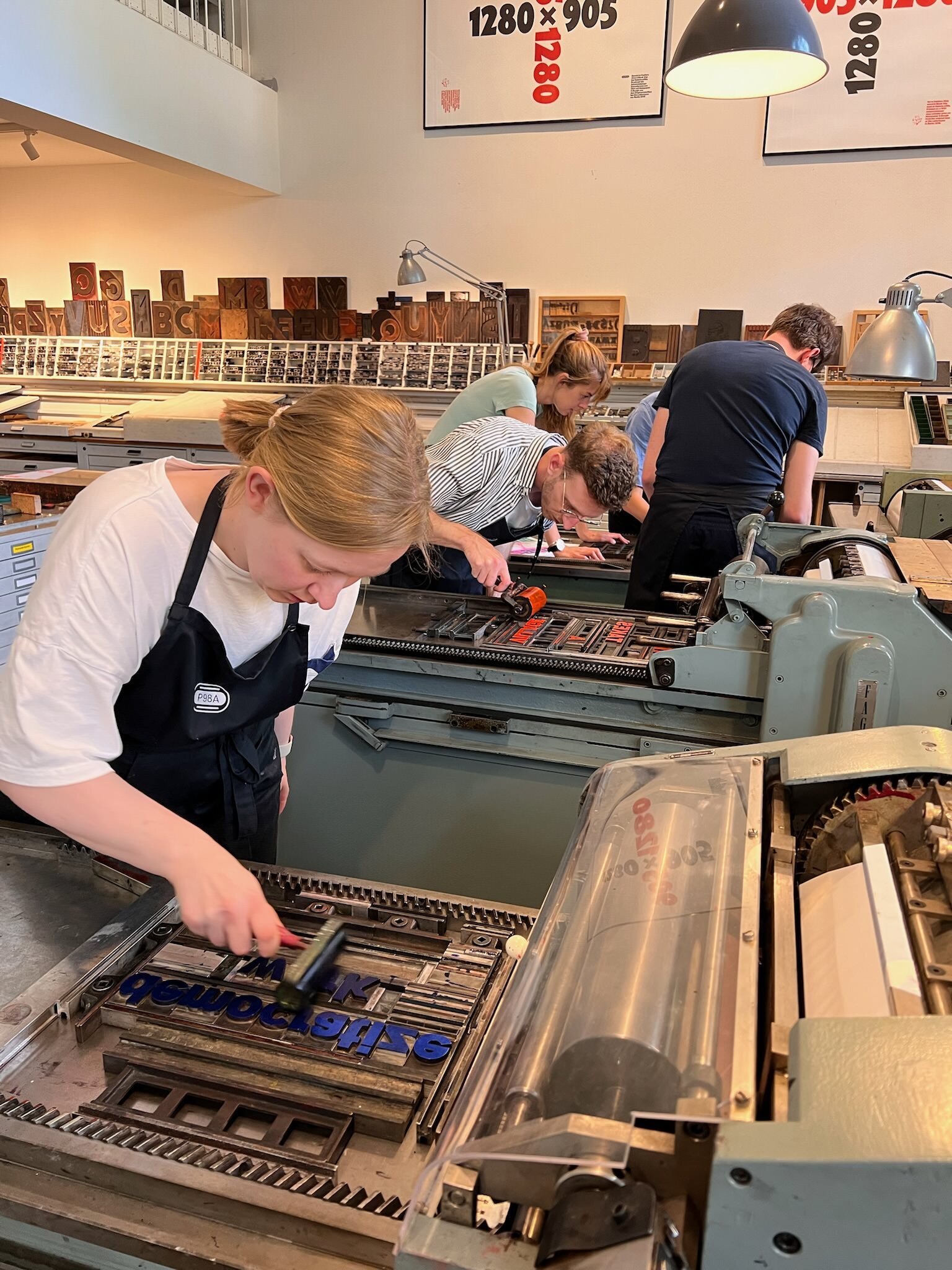 Photo of several letterpress machines and people standing around them, applying paint, for printing poster, everyone appears very focused