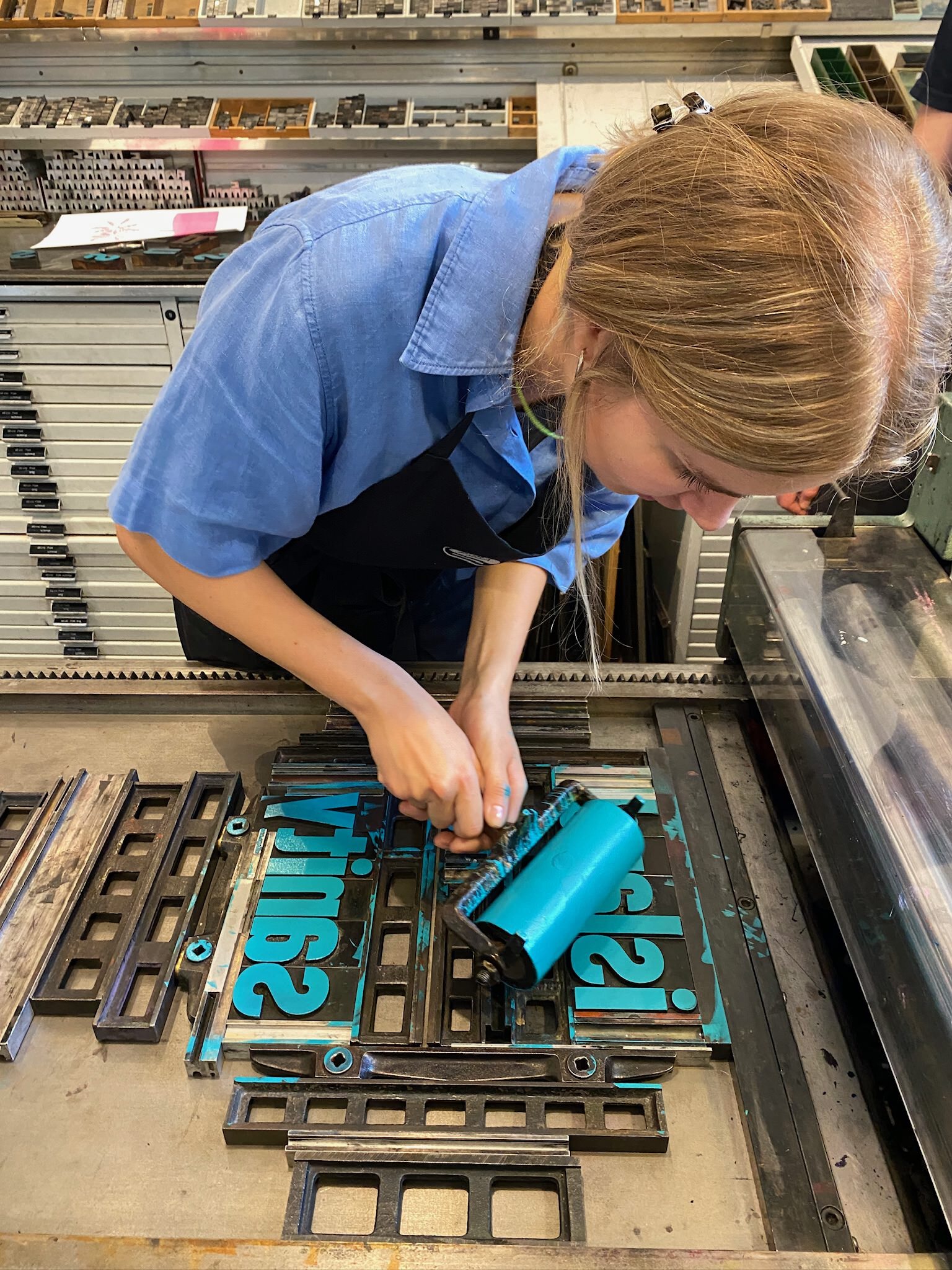 Photo of a woman applying paint with a roller to metal type arranged inside a printing machine