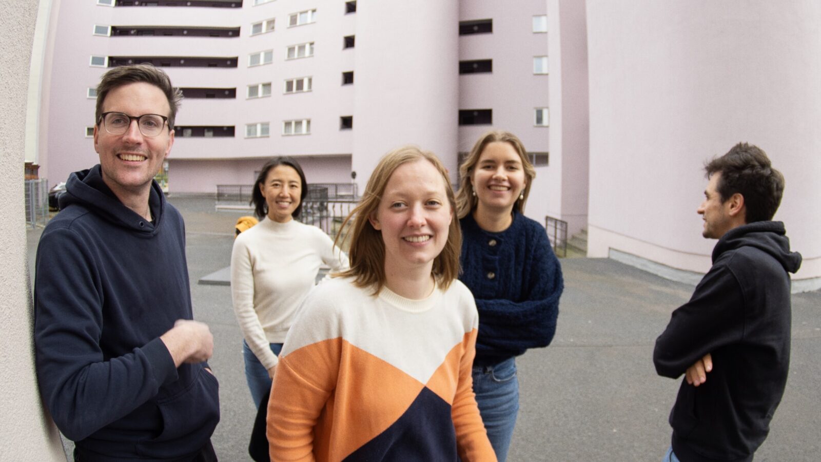 Fisheye photo of the Village One Team, standing in a brutalist Berlin backyard, five people visible in total (three women, two men), four of them looking and smiling at the camera, one person only visible from the side