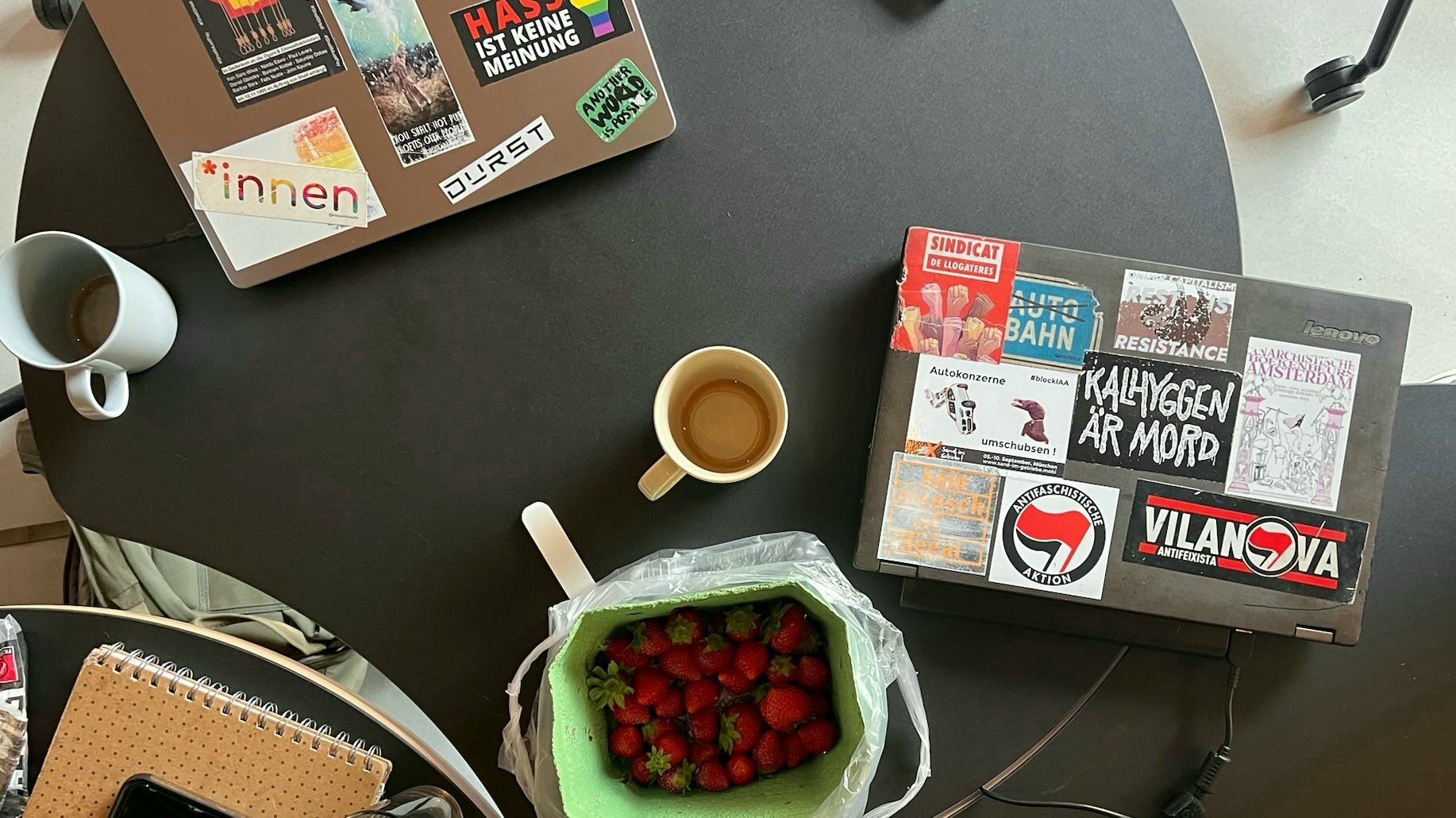 Photo of a meeting room desk, with strawberries, coffee cups, a notebook and two laptops visible. The laptops feature countless stickers, with many progressive slogans.