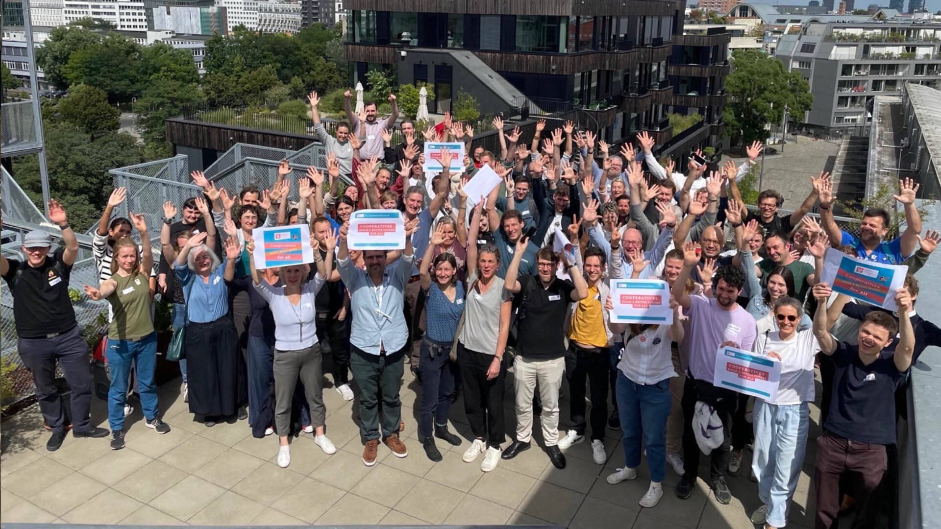 Photo of a large crowd of people, standing on a roof terrace in summer, cheering at the camera and holding up signs that read “Cooperatives build a better world for all”