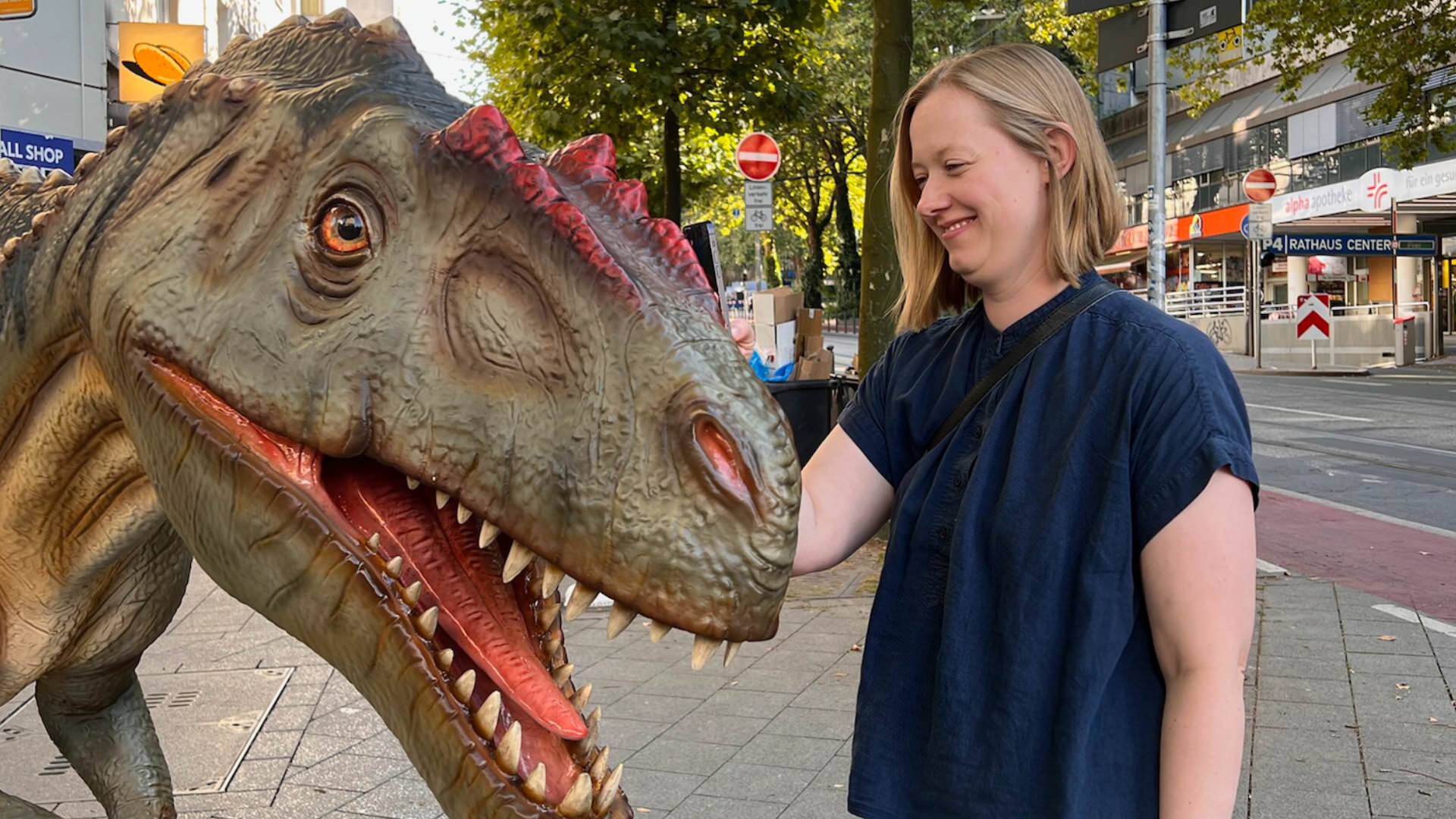 A woman standing in a sunny street next to a life-sized dinosaur sculpture, smiling