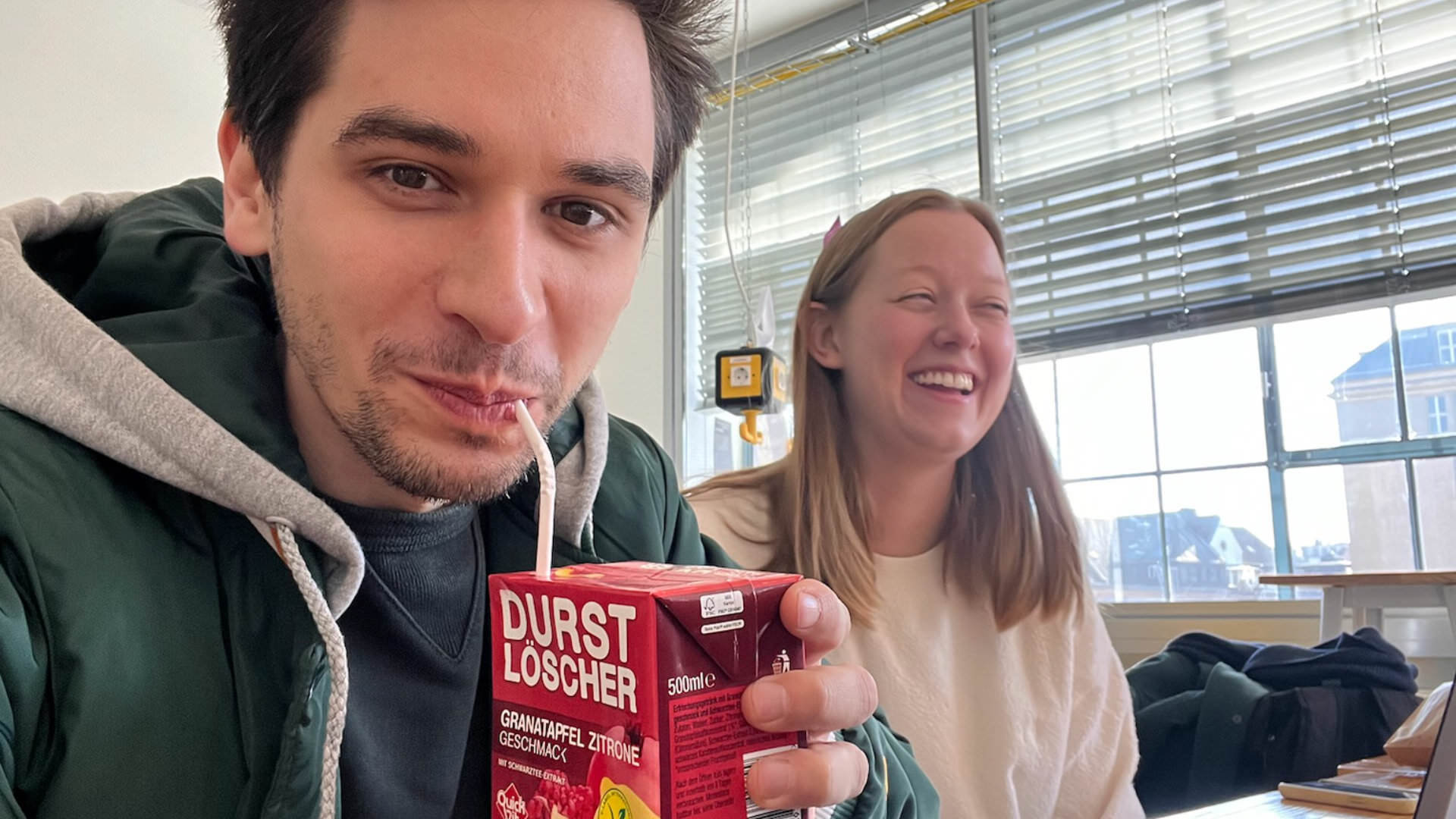 Photo of a man and a woman looking at the camera – the former drinking a “Durstlöscher” drink through a straw, with the woman laughing, sitting in a bright room