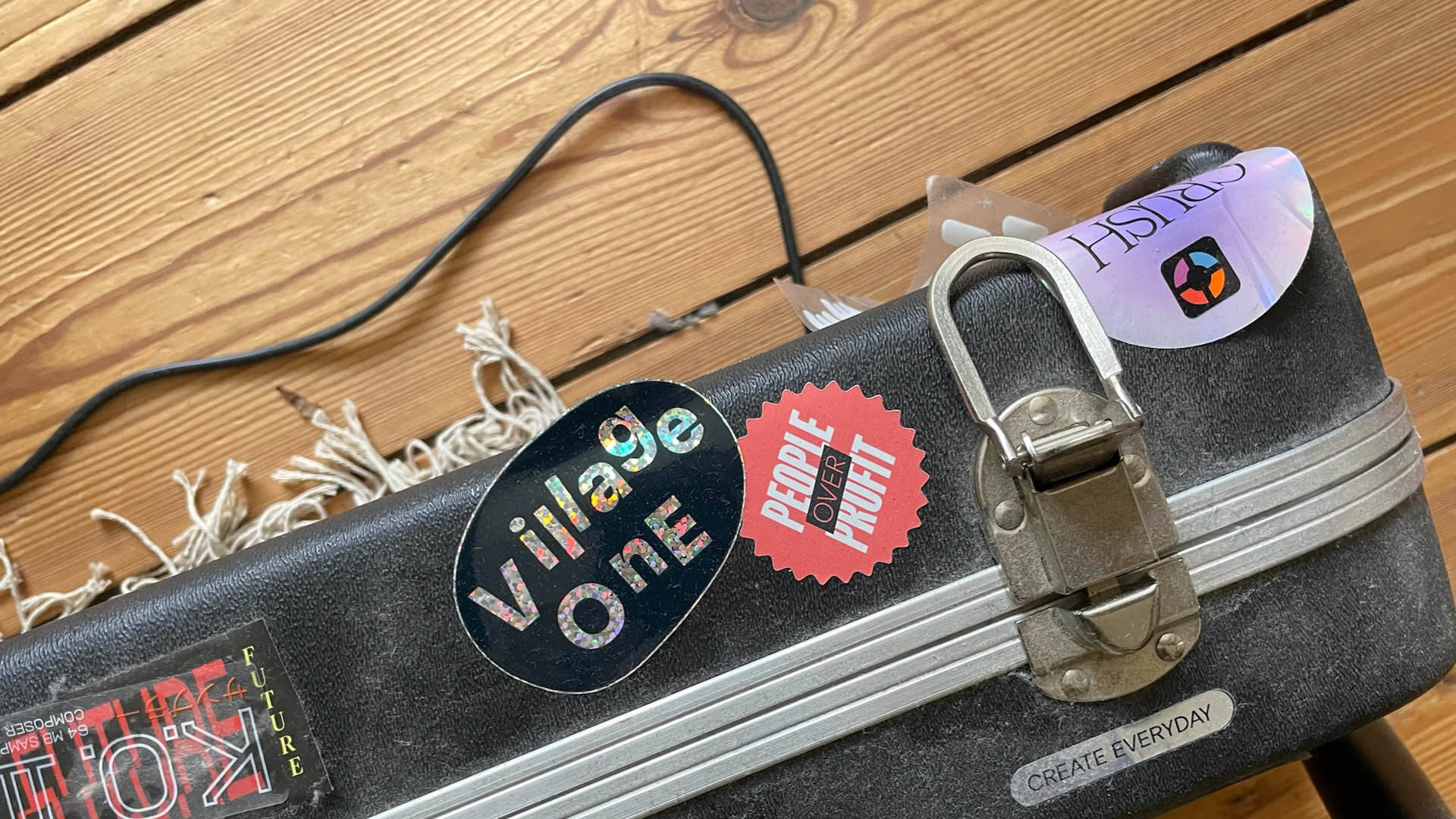 Photo of a beat-up suitcase with several stickers on it, two of them saying “Village One” and “People over Profit”, wooden floor below