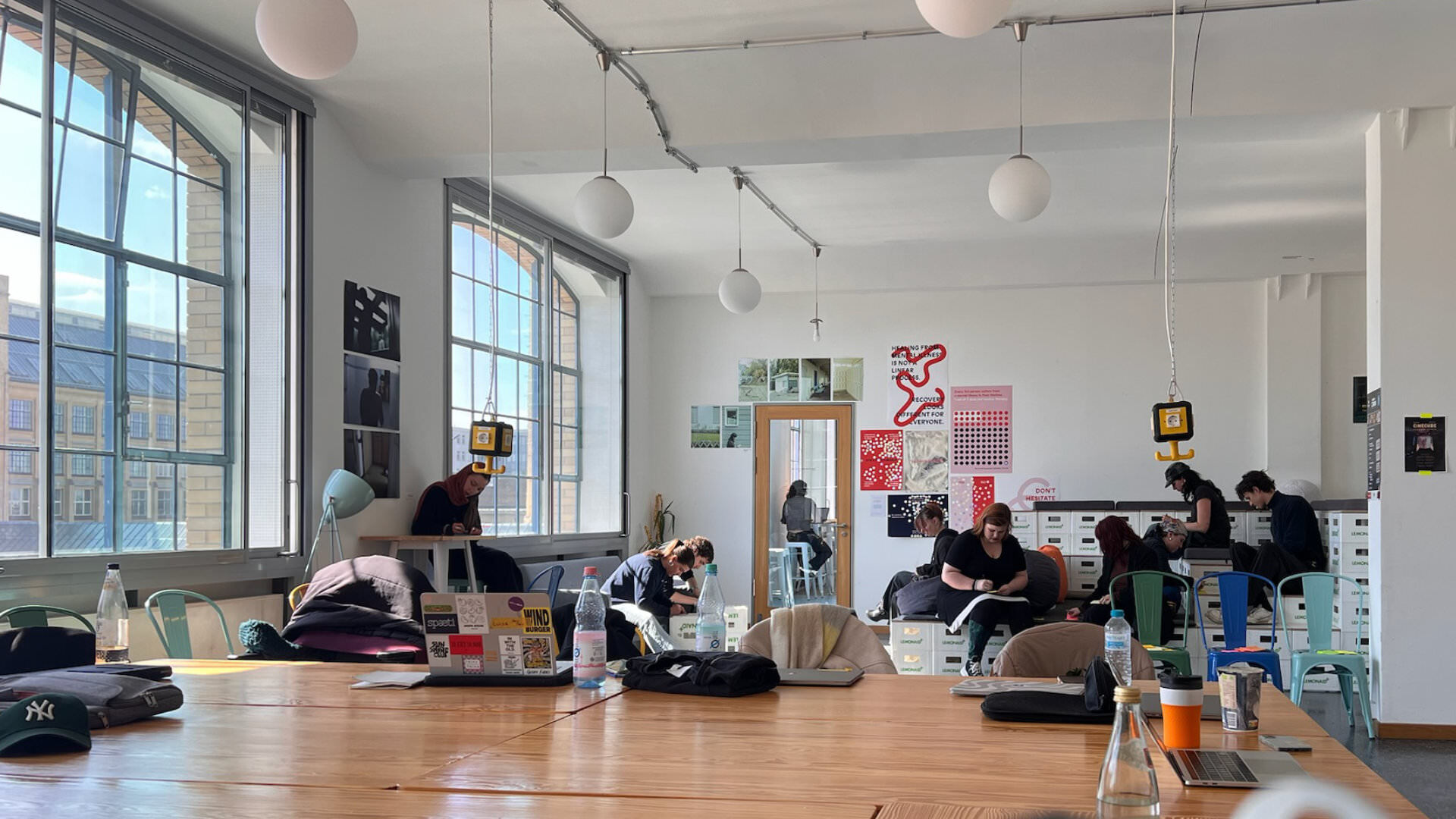 Photo of a large workshop room in an industrial building, very bright, with large windows and approx. 10 people sitting around across the room, studying and writing. Table in front, with laptops, cups and bottles.