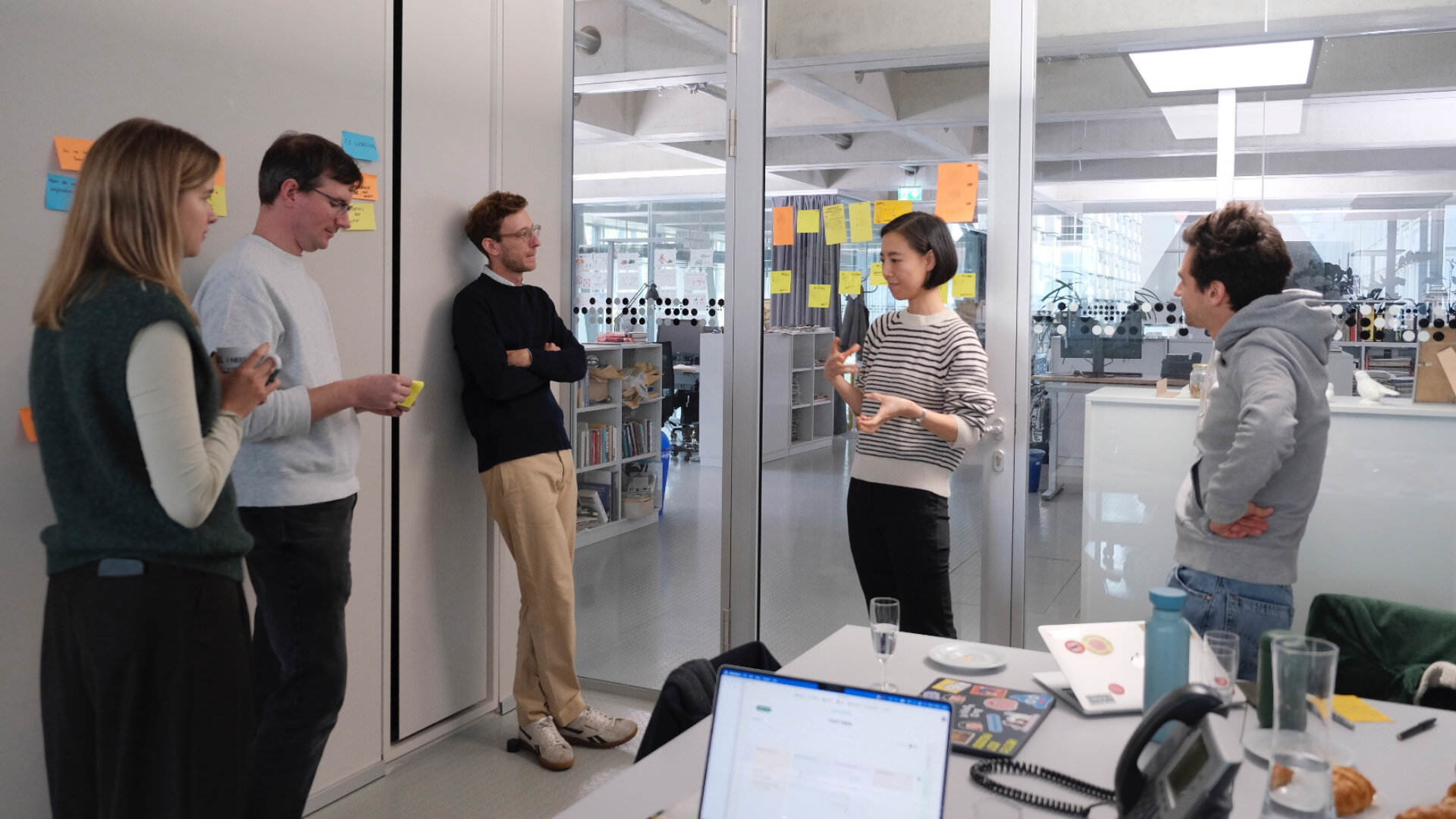 Photo of a team of five people standing in a half circle in an office setting, discussing and pondering a heavy topic