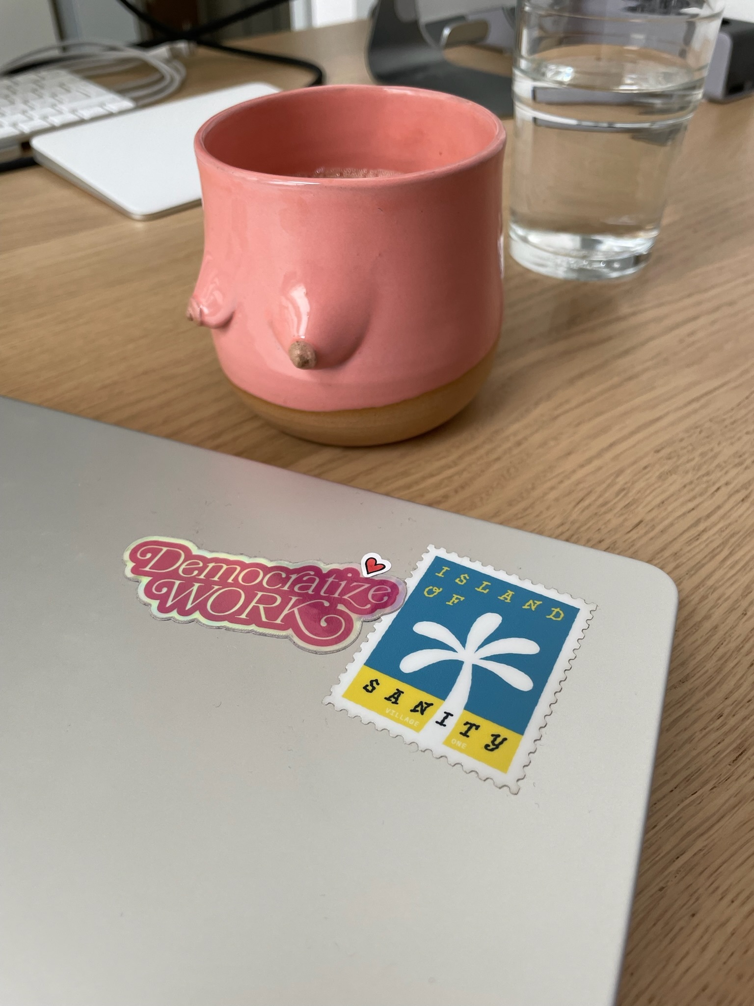 A photo of a desk situation with a laptop lid visible in the foreground, decorated with two stickers that say “Democratize Work” and “Island of Sanity”, and a coffee mug and water glass visible in the background