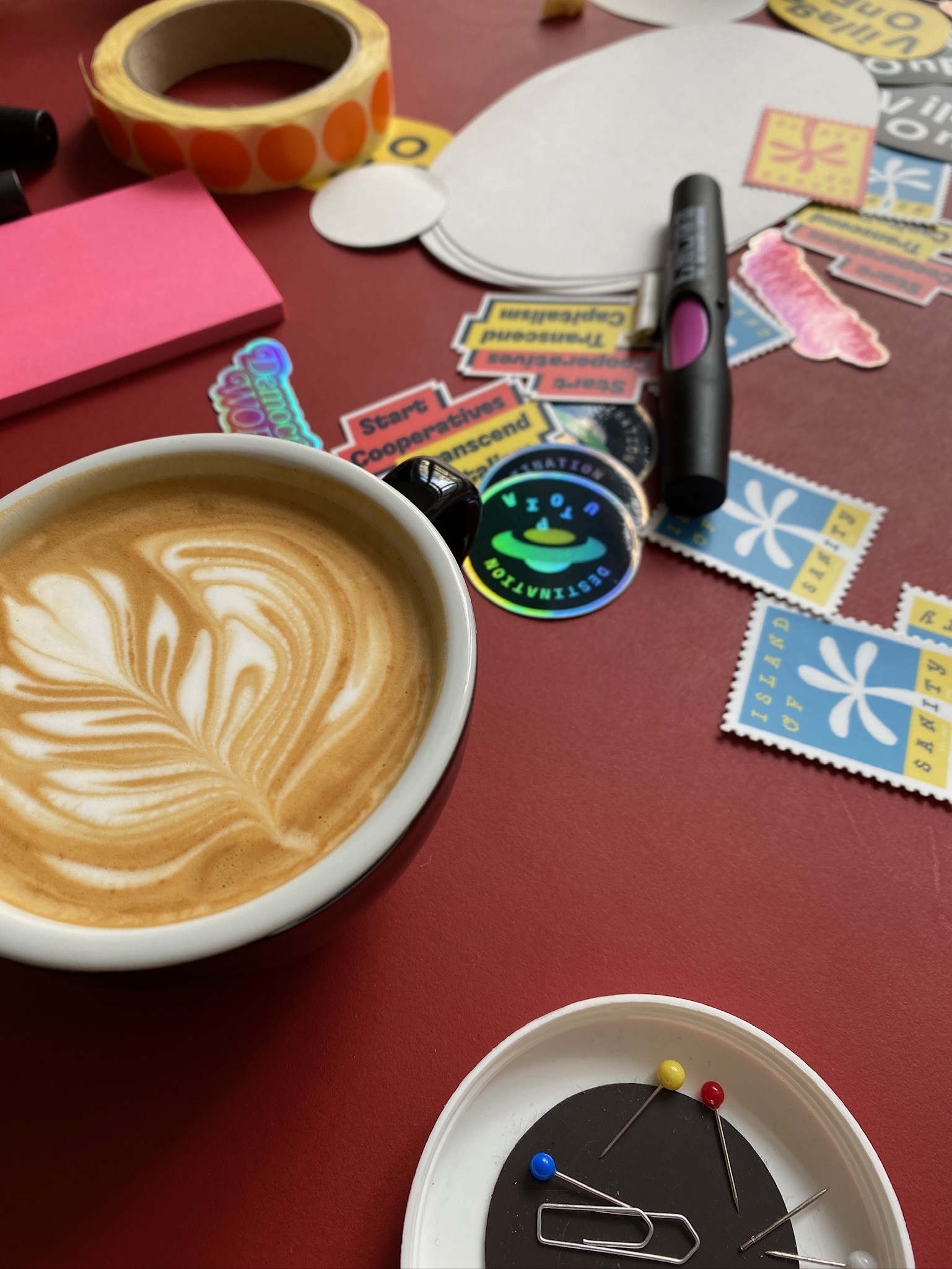 Photo of a desk, with a cup of coffee, marker pens and a bunch of stickers, with messages such as “Start Cooperatives, Transcend Capitalism”, “Destination Utopia” or “Island of Sanity”