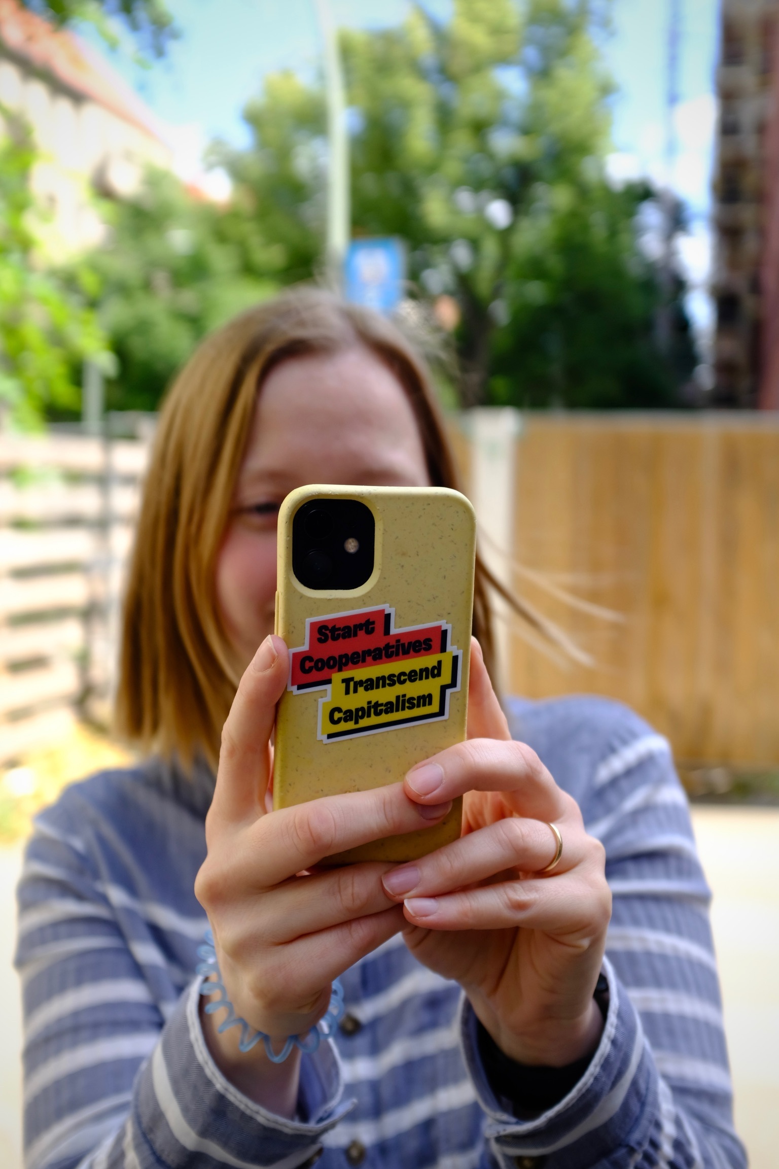 Photo of a blonde woman holding up her phone into the camera, in a yellow case with a sticker that says “Start Cooperatives, Transcend Capitalism”