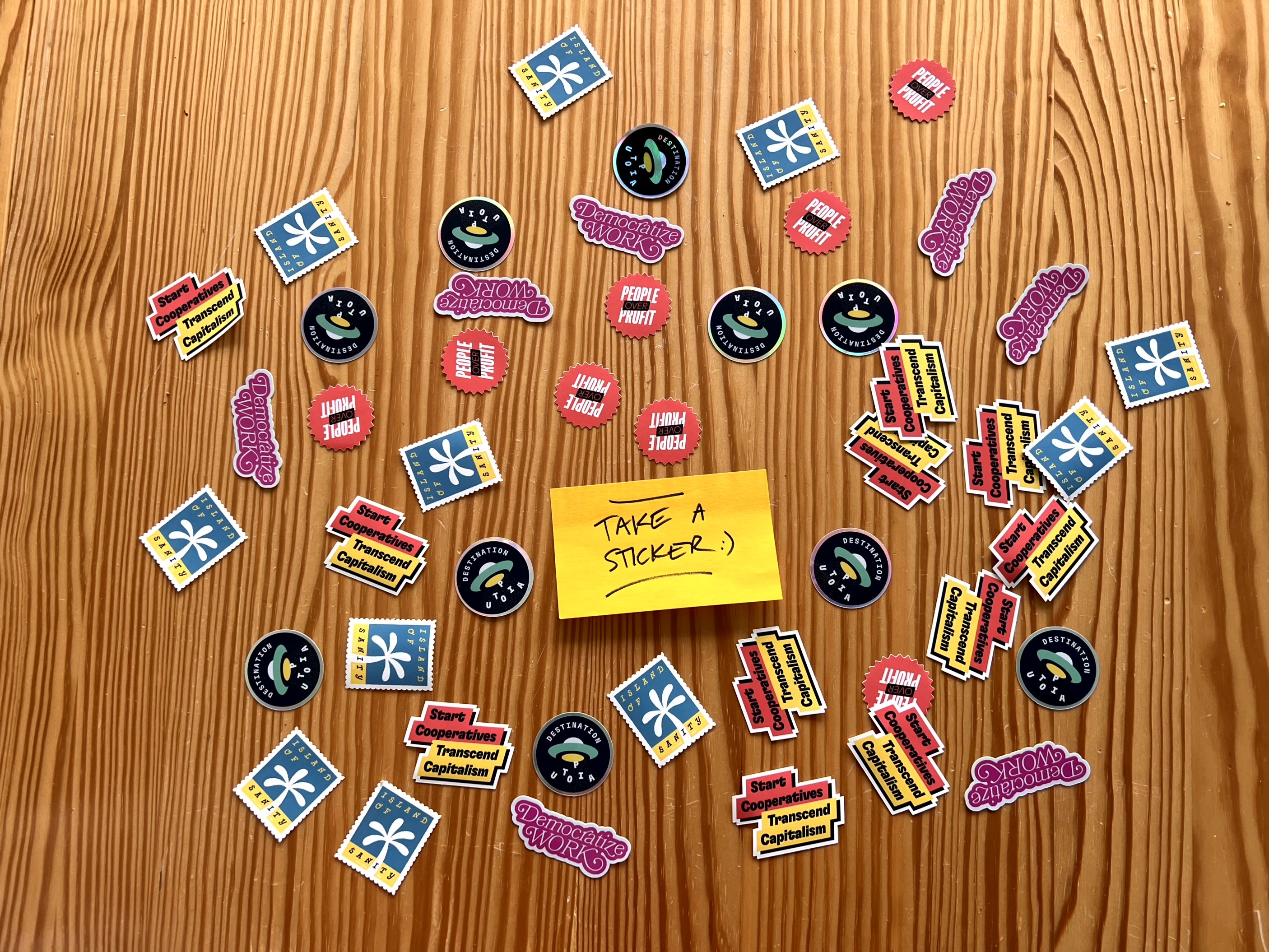 Photo of a wooden desk, with a sticky note in the center that says “Take a sticker! :)” and lots of stickers arranged around it, with messages such as “Start Cooperatives, Transcend Capitalism”, “People over Profit”, “Destination Utopia” or “Island of Sanity”