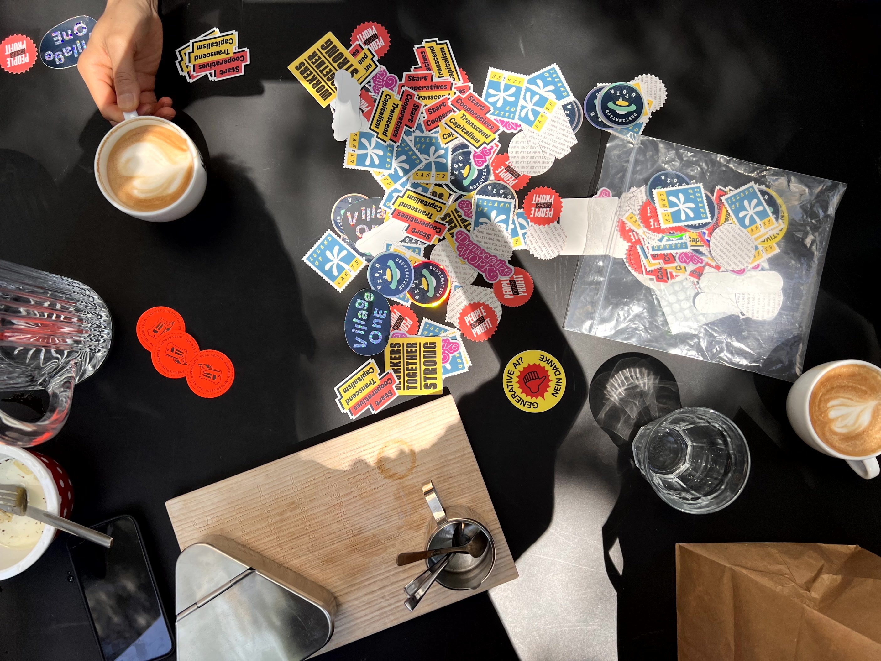 Photo of a desk, with cups of coffee, some kitchen utensils and a bunch of stickers, with messages such as “Start Cooperatives, Transcend Capitalism”, “Destination Utopia”, “Democratize Work” or “Island of Sanity”