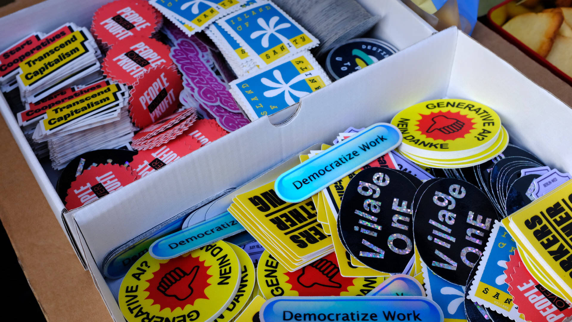 Photo of a box of colorful stickers, saying “Democratize Work”, “Village One”, “People over Profit”, “Start Cooperatives, Transcend Capitalism” and “Generative AI? Nein Danke”